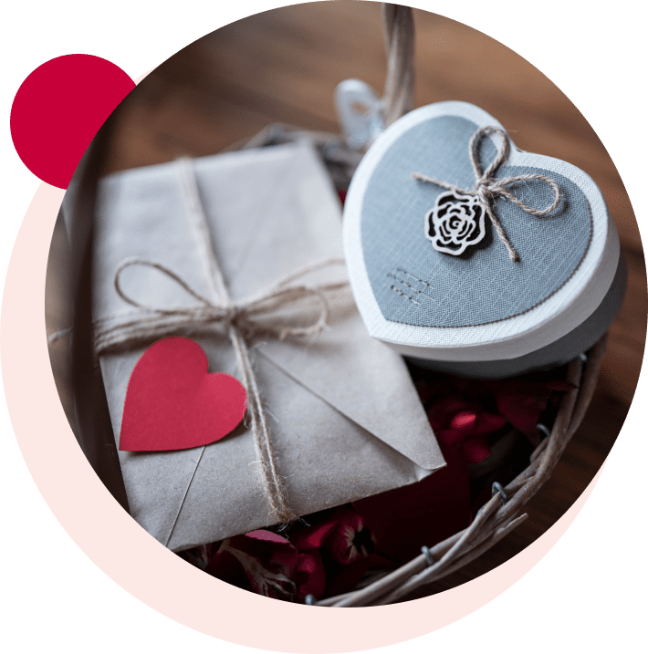 A basket contains a brown paper-wrapped gift tied with twine, a red paper heart, and a heart-shaped box with a fabric cover and floral detail.